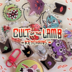 May include: A collection of clear acrylic keychains featuring various cartoon characters and designs. The keychains include a green monster, a purple ghost, and a blue creature. The text "CULT OF THE LAMB" and "#KEYCHANS#" is visible. A pink carabiner is also present.