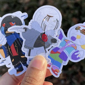 Three skeletons. The one on the left is a black skeleton is string on his hands, the one in the middle is shrugging as he holds a knife, and the one on the left is holding his hands out in upside down peace signs.