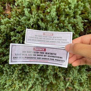 A rectangle die cut sticker of a warning sign with the message NOTICE, DO NOT FEED THE MACHINE, LEST OUR ART BE DONE BY ALGORITHMS AND OUR PASSIONS SQUASHED FOR PROFITS.