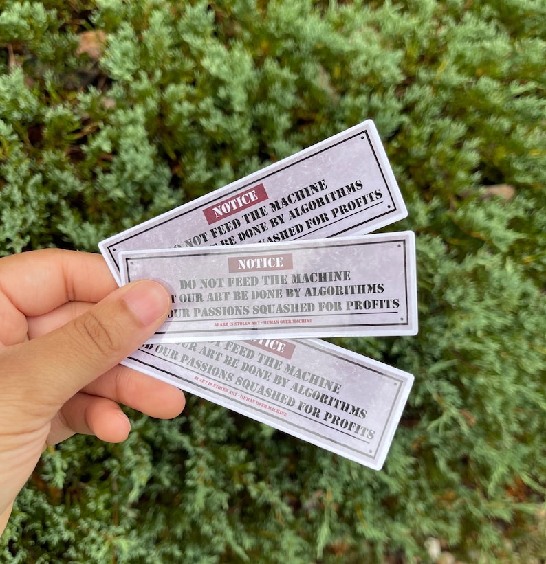 A rectangle die cut sticker of a warning sign with the message NOTICE, DO NOT FEED THE MACHINE, LEST OUR ART BE DONE BY ALGORITHMS AND OUR PASSIONS SQUASHED FOR PROFITS.