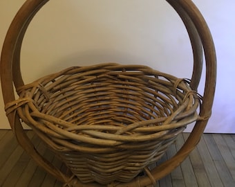 May include: A woven wicker basket with a large, round handle. The basket is made of natural brown wicker and has a slightly conical shape.