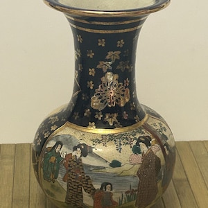 May include: A navy blue ceramic vase with a wide base and a flared rim. The vase features a painted scene of figures in traditional Japanese attire, with gold floral accents and trim. The vase is approximately 8 inches tall.