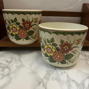 May include: Two white ceramic flower pots with a green, yellow, red, and brown floral pattern. The pots are on a marble surface.