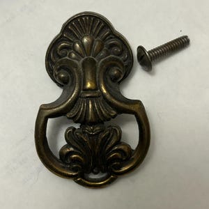 May include: Ornate antique brass drawer pull with a decorative floral design and a matching screw. The pull features a curved handle and detailed embellishments, perfect for vintage furniture restoration projects.