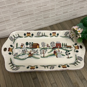 May include: A white ceramic serving tray with a brown rim and a green, red, and yellow country scene with houses, trees, and people. The tray has two handles on the sides.