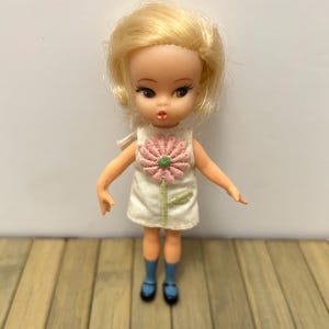 May include: A vintage doll with blonde hair, wearing a white dress with a pink flower applique. The doll has blue socks and black shoes. The doll is standing on a wooden surface.