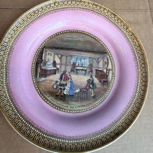 May include: A round ceramic plate with a pink border and a gold-trimmed rim featuring a decorative pattern. The center displays a detailed illustration of an interior room with figures and the text: THE ROOM IN WHICH SHAKSPEARE WAS BORN 1564 STRATFORD ON AVON.