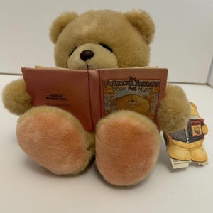 May include: A beige plush teddy bear wearing a red jacket is sitting and reading a pink book titled "Forever Friends Book for Mums".