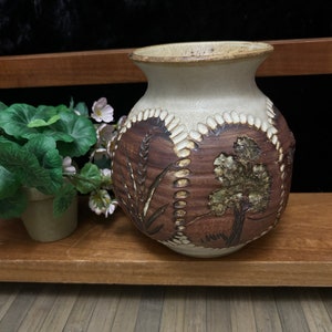 May include: A white ceramic vase with a brown and green floral design. The vase has a rounded body and a wide mouth.