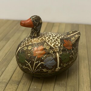 May include: Ornamental duck figurine featuring detailed floral and geometric designs. The duck has a brown body with orange, green, and blue leaf accents. The beak is orange. The duck is on a wooden surface.