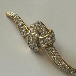 May include: A gold-toned brooch featuring a knot design. The knot and the curved bands are encrusted with clear, sparkling rhinestones. The brooch has a classic, elegant style, suitable for formal wear.