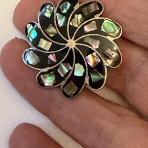 May include: A flower-shaped brooch with a silver-tone border. The brooch features black enamel petals inlaid with iridescent abalone shell pieces. The center of the brooch has a silver-tone, star-like design.