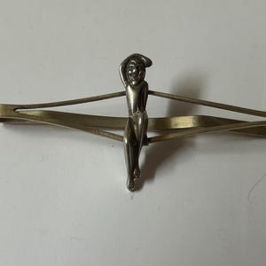 May include: A vintage tie clip with a silver-toned figure of a person. The figure is centered on a gold-toned bar clip. The person is wearing a hat and has their arm raised. The clip is designed to hold a tie in place.
