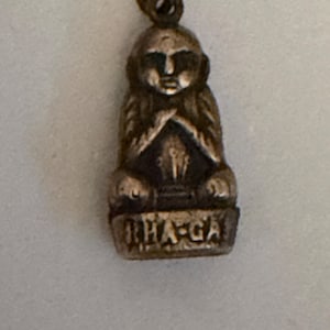 May include: A small, antique-looking metal pendant in the shape of a seated figure with crossed arms. The pendant has a loop at the top for a chain and the word "THA-GA" is inscribed on the base. The pendant has a tarnished, aged appearance.