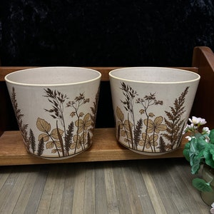 May include: Two white ceramic plant pots with a brown floral design. The pots are sitting on a wooden shelf.