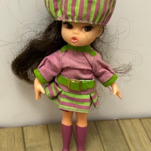 Peut inclure: Poupée vintage vêtue d'une robe rose, d'un chapeau rayé vert et rose et d'une jupe assortie. La poupée a des cheveux bruns, des yeux bruns et porte des bottes hautes violettes et des chaussures marron. La tenue comprend une ceinture verte avec une boucle dorée.