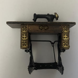 May include: A miniature wooden sewing machine with a black metal base and a black metal sewing machine on top. The sewing machine has gold accents and a wooden top.