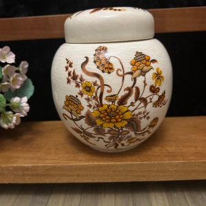 以下が含まれることがあります： クリーム色のセラミックジャーで、蓋付き、黄色と茶色の花柄で飾られています。ジャーは丸い形で、木製の棚の上にあります。花柄のデザインはジャーの表面を覆っています。