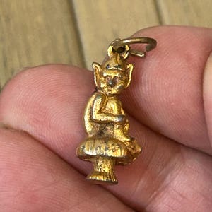 Peut inclure: Un petit charme doré représentant une figurine d'elfe ou de gnome assis. Le charme a une boucle en haut pour l'attacher et est assis sur une base en forme de champignon. Les détails de la figurine sont visibles, avec des oreilles pointues et un petit chapeau.