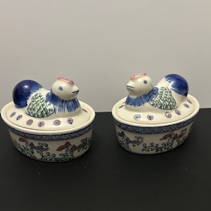 以下が含まれることがあります： アヒルの形をしたセラミック製の蓋付きの器が2つ。アヒルは青、緑、白のディテール。ベースは楕円形で、青と赤の花柄。蓋には青い鶏冠とピンクと白の頭が付いています。