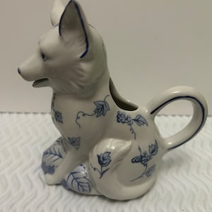 Puede incluir: Una jarra de crema de cerámica blanca con forma de zorro con diseños florales azules. La jarra tiene un pico y un asa.