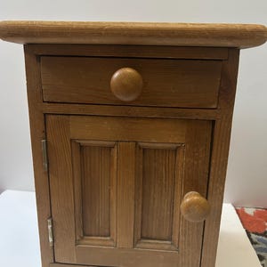 May include: A small wooden cabinet with a drawer and a door. The cabinet is made of light brown wood and has a rounded top. The drawer and door each have a round wooden knob. The cabinet is a rustic style.