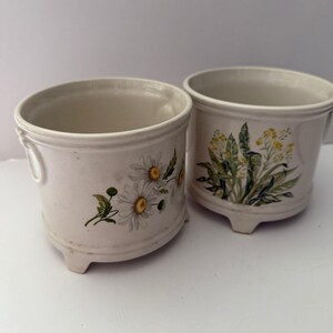 Peut inclure: Deux pots de fleurs en céramique blanc cassé avec des motifs floraux. L'un présente des marguerites, l'autre des fleurs jaunes et du feuillage vert. Chaque pot a une forme arrondie, de petits pieds et un bord décoratif.