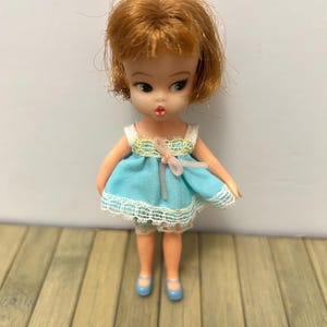 Peut inclure: Une poupée vintage aux cheveux roux, vêtue d'une robe bleu clair avec une bordure en dentelle blanche et un nœud rose. La poupée porte des chaussures bleues et est posée sur une surface en bois.