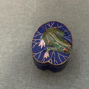 May include: A small, oval-shaped trinket box with a detailed frog design. The frog is green with red eyes, resting on a blue lily pad with gold accents. The box has a dark blue base and gold trim.