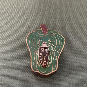 May include: A decorative pin in the shape of a green pepper with a gold outline. The pepper has a brown stem and a brown and black insect in the centre. The pin is on a green textured surface.