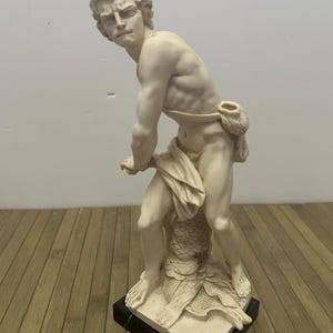 以下が含まれることがあります： 筋肉質な男性像のクリーム色の像。決意した表情で前に傾いている。像は白黒の台座に取り付けられています。この像は布で覆われており、質感のある台座の上に立っています。