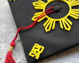 May include: Black graduation cap with yellow accents, including a sunburst design, a star, and the year "2024". A red tassel hangs from the cap. The cap is a symbol of academic achievement and celebration.