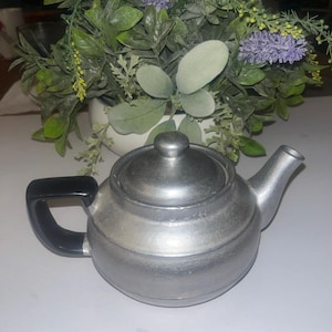 May include: A small, silver-colored teapot with a black handle and a lid. The teapot has a rounded body and a spout. The background features a white pot with green and purple faux flowers.