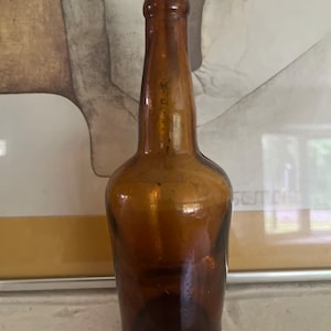 May include: A vintage amber glass bottle with a long neck and rounded body. The bottle has a dark brown hue and appears to be empty. The bottle is likely an antique or collectible item.