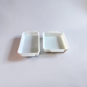 May include: Two white rectangular baking dishes with rounded corners and small handles. The dishes are empty and sit side-by-side on a white surface. The dishes are made of a glossy material.