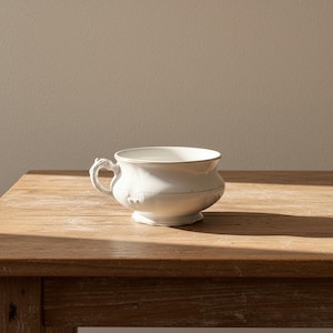May include: A white ceramic bowl with a handle and a decorative rim. The bowl is sitting on a wooden table. The handle has a unique, ornate design. The bowl is approximately 6 inches in diameter.