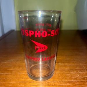 May include: A clear glass measuring cup with red lettering that reads "Fleet Phospho-Soda Buffered Laxative".