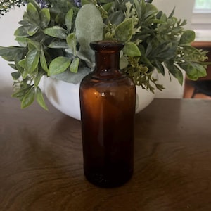 May include: A small, brown glass bottle with a rounded shoulder and a narrow neck. The bottle is cylindrical and has a dark amber color. It is displayed on a wooden surface, with greenery in the background.