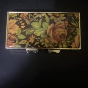 May include: A small, rectangular metal box with a hinged lid. The box is decorated with a floral pattern in shades of brown, yellow, and orange. The flowers include roses, daisies, and other small blooms.