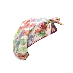 May include: A white fabric shower cap with a pink trim and a pineapple print. The cap has a tie at the top and a label that reads "Soft as silk".