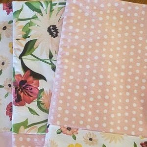 May include: Set of four pink and white kitchen towels. Two towels feature a floral pattern with pink, red, and yellow flowers. One towel has a white polka dot pattern on a pink background. The fourth towel has a white floral pattern with yellow and pink flowers on a pink background.