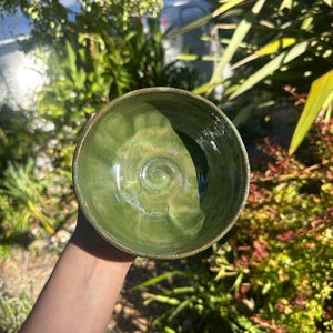 May include: A handmade ceramic bowl with a glossy green glaze. The bowl has a rounded shape with a slightly textured interior. The rim of the bowl has a darker, earthy tone. The bowl is held up against a background of green foliage.