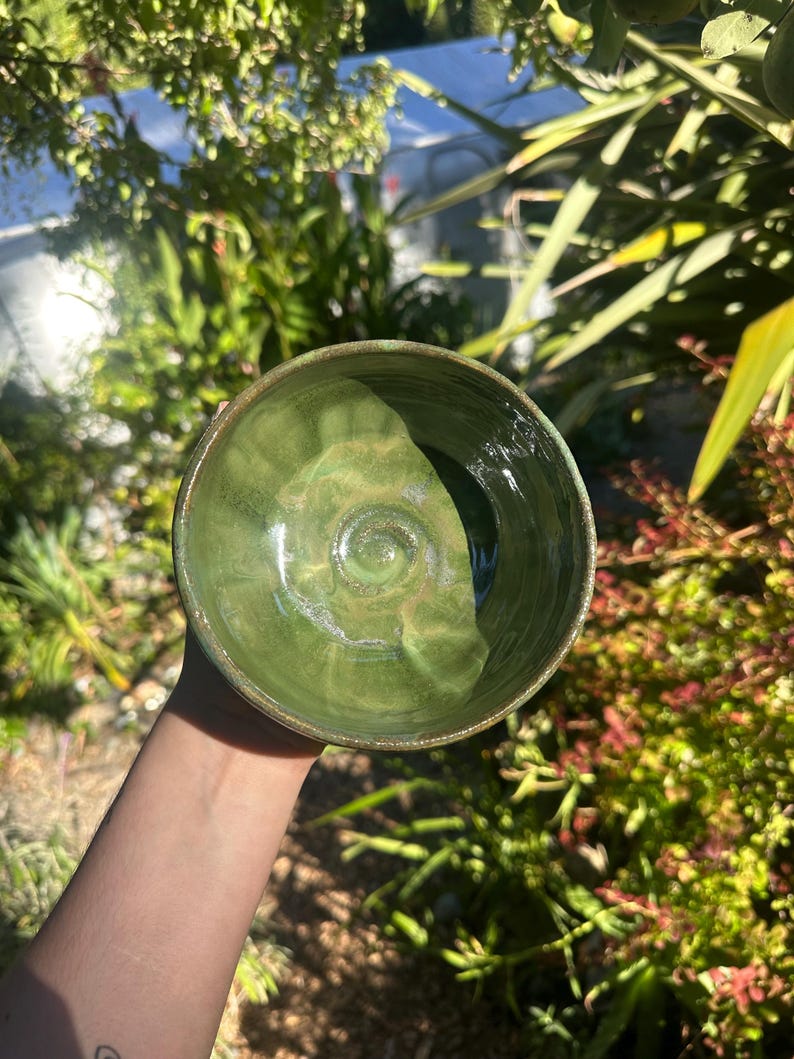 May include: A handmade ceramic bowl with a glossy green glaze. The bowl has a rounded shape with a slightly textured interior. The rim of the bowl has a darker, earthy tone. The bowl is held up against a background of green foliage.