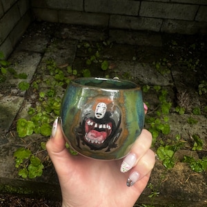 May include: Hand-painted ceramic mug with a glossy, teal and green glaze. The mug features a detailed illustration of a character with a wide open mouth and a white mask. The mug is round and appears to be handmade.