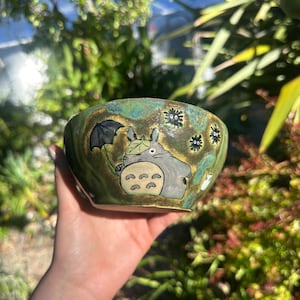 May include: Hand-painted ceramic bowl with a green and gold glaze. The bowl features a cartoon illustration of Totoro holding an umbrella, with soot sprites around the rim. The bowl is held in a hand.