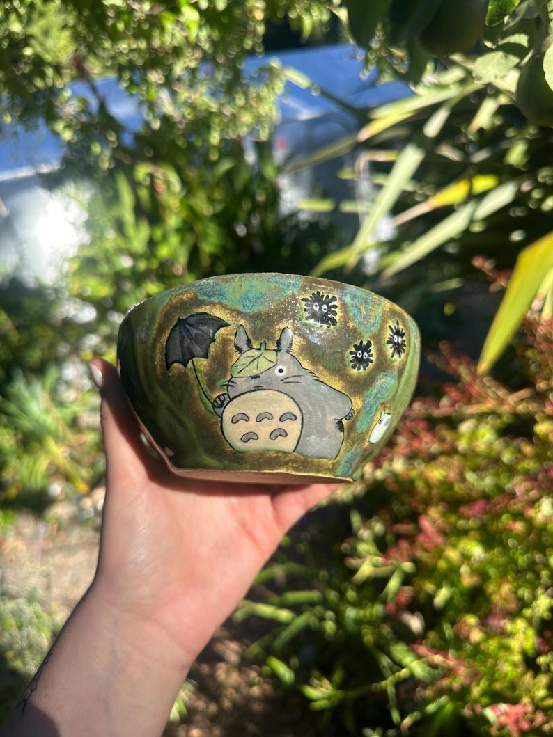 May include: Hand-painted ceramic bowl with a green and gold glaze. The bowl features a cartoon illustration of Totoro holding an umbrella, with soot sprites around the rim. The bowl is held in a hand.