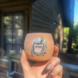 May include: A small, round, terracotta-colored ceramic pot. The pot features a hand-painted illustration of a character from the animated film "Spirited Away." The character is depicted in shades of gray and white. The pot is held in a hand.