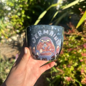May include: A hand-painted ceramic cup with a dark blue exterior. The cup features the name "Jermaine" in white lettering and a cartoon illustration of a character. The background is a mix of purple and blue.