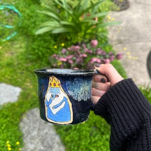 May include: A dark blue ceramic mug with a cartoon image of a character wearing a crown and blue robe. The mug has a black handle and a textured, glazed finish. The background is blurred with green foliage.