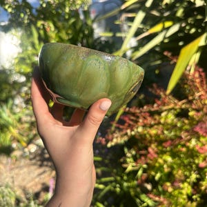 May include: A handmade, green ceramic bowl with a textured surface and a glossy finish. The bowl has a unique, organic shape and a dark brown rim. The bowl is held in a hand against a blurred background of green foliage.